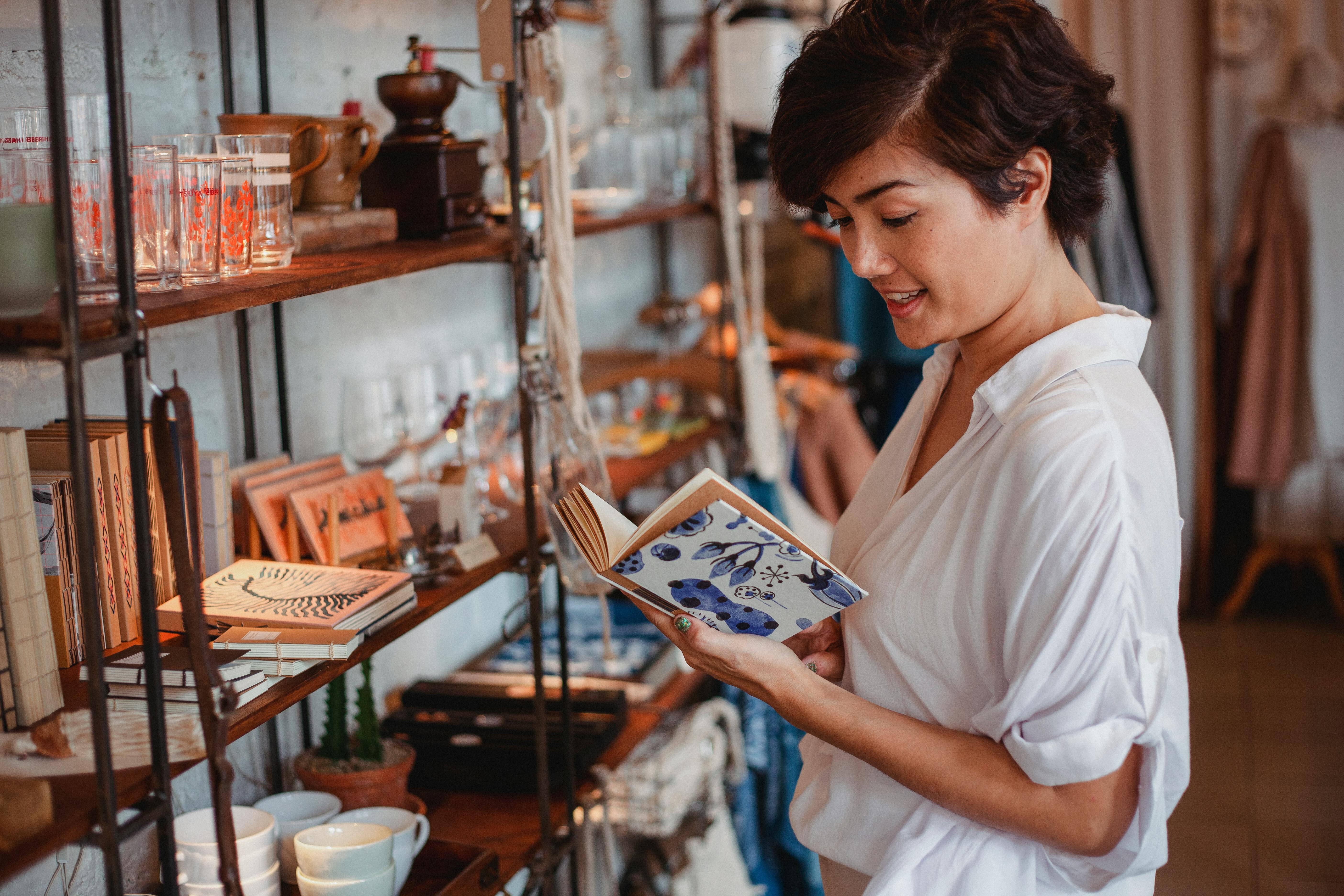 Asian woman in a boutique reading a book among stylish home decor items on shelves.